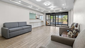 Une pièce spacieuse avec un canapé gris, un fauteuil plus petit, un plancher en bois, un coin cuisine et une porte-fenêtre coulissante donnant sur un espace extérieur.
