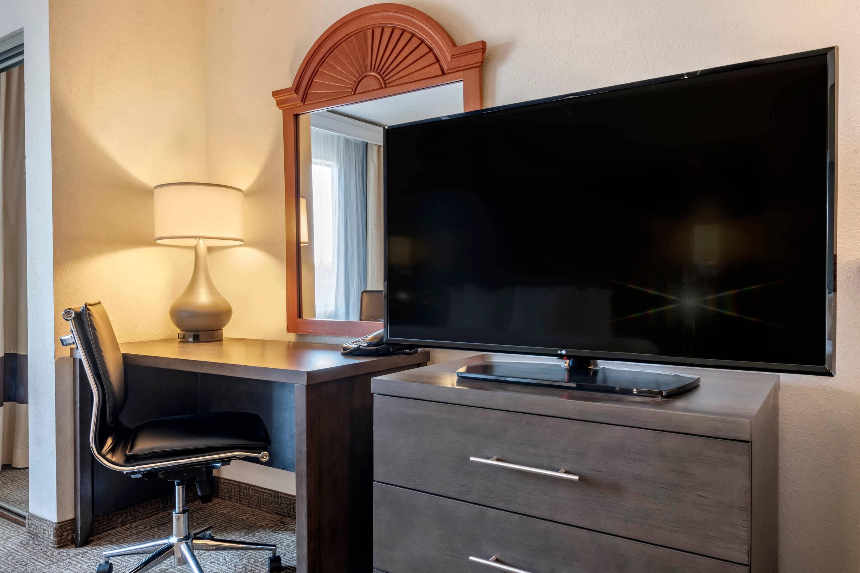 Habitación de hotel con una televisión de pantalla plana sobre un tocador, un escritorio con una silla y una lámpara.