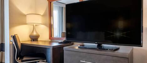 Habitación de hotel con una televisión de pantalla plana sobre un tocador, un escritorio con una silla y una lámpara.