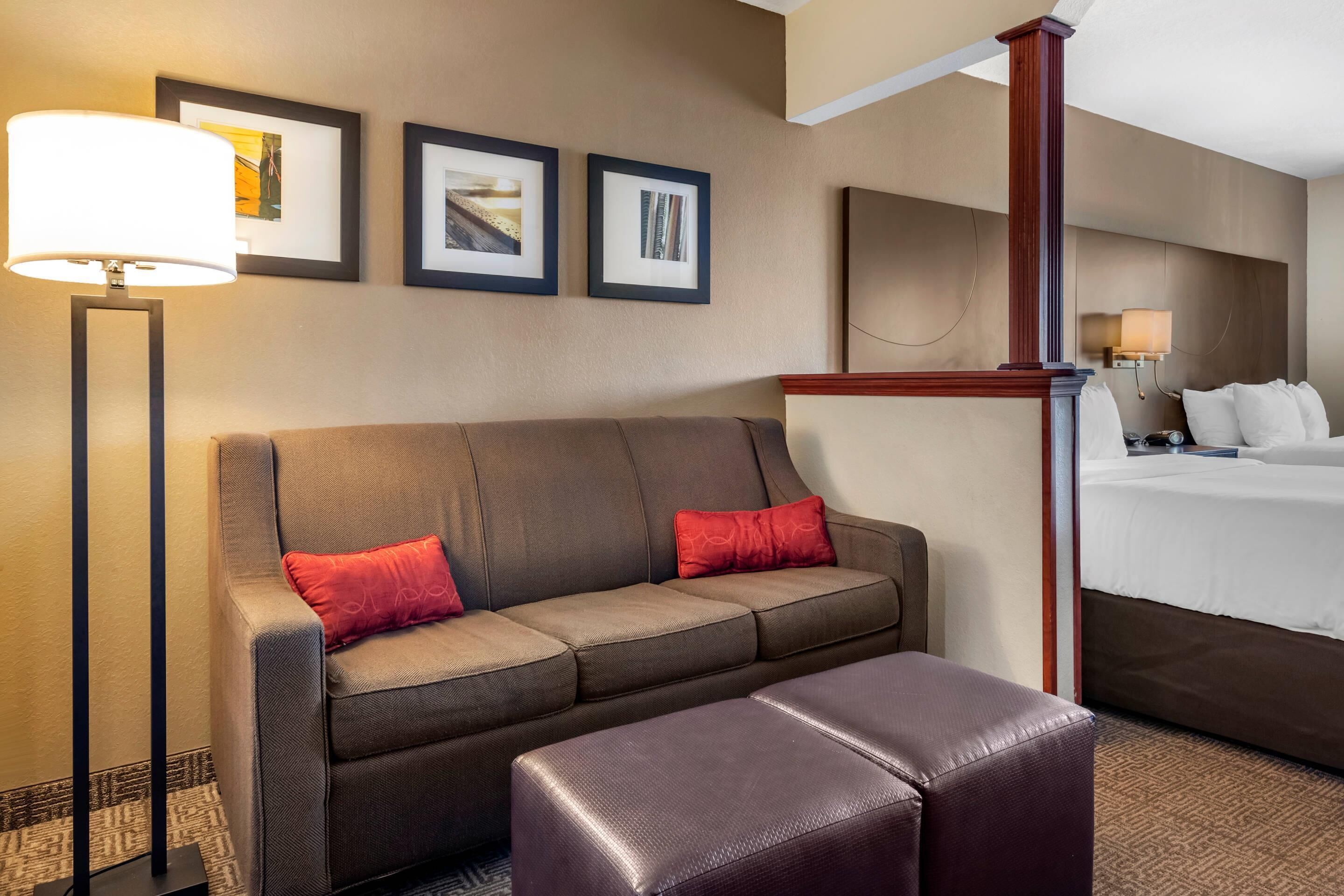 A hotel room with a brown sofa, two red pillows, a wooden headboard, a bed with white linens, and a floor lamp.
