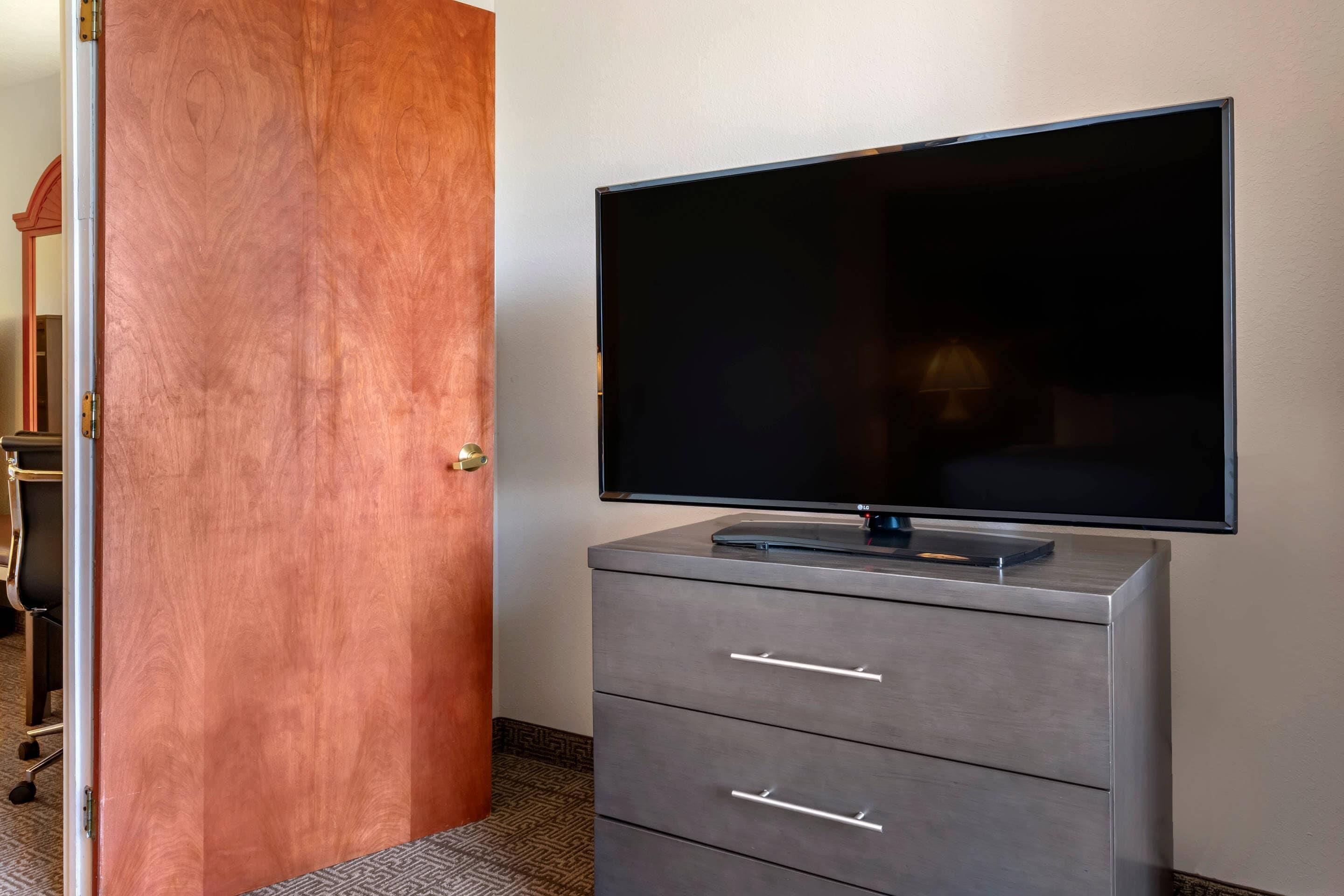 A hotel room with a flat-screen TV on a grey dresser, a wooden door, and a patterned carpet.