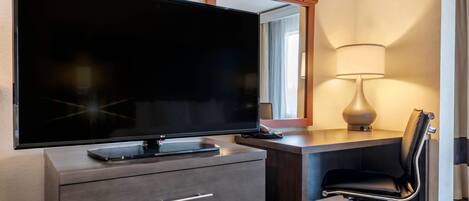 Quarto de hotel com televisão de tela plana sobre uma cômoda de madeira, uma escrivaninha com cadeira, luminária e espelho acima da cômoda.