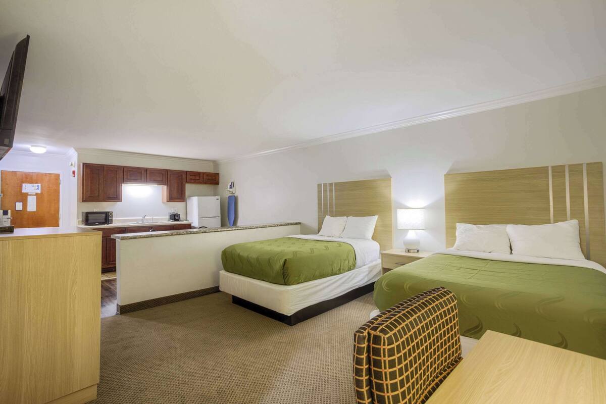 A hotel room with two beds, a kitchenette, a TV, and a small seating area.