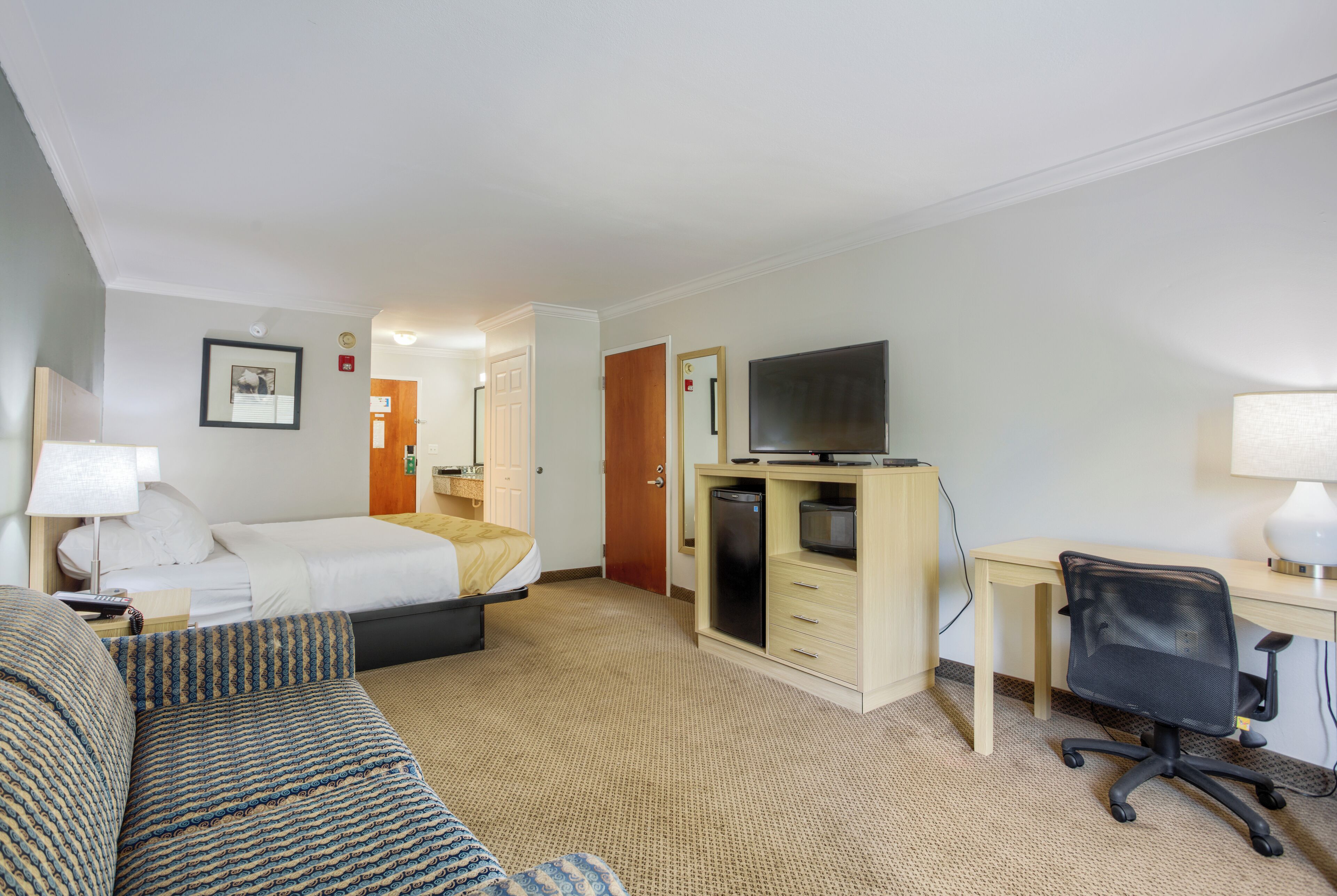 Habitación de hotel con cama, escritorio con computadora, televisor, sofá y una pequeña kitchenette.