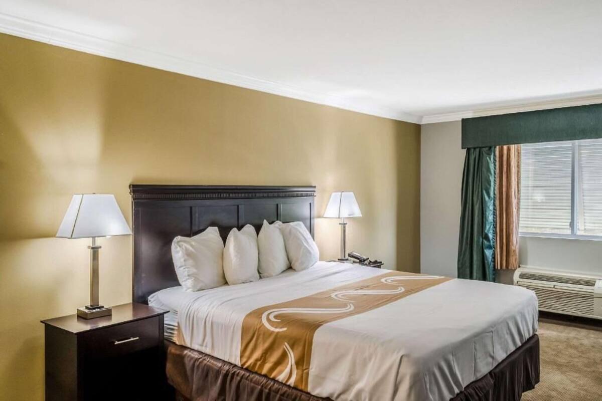 A hotel room with a large bed, two bedside tables with lamps, a window with curtains, and a wall-mounted air conditioning unit.