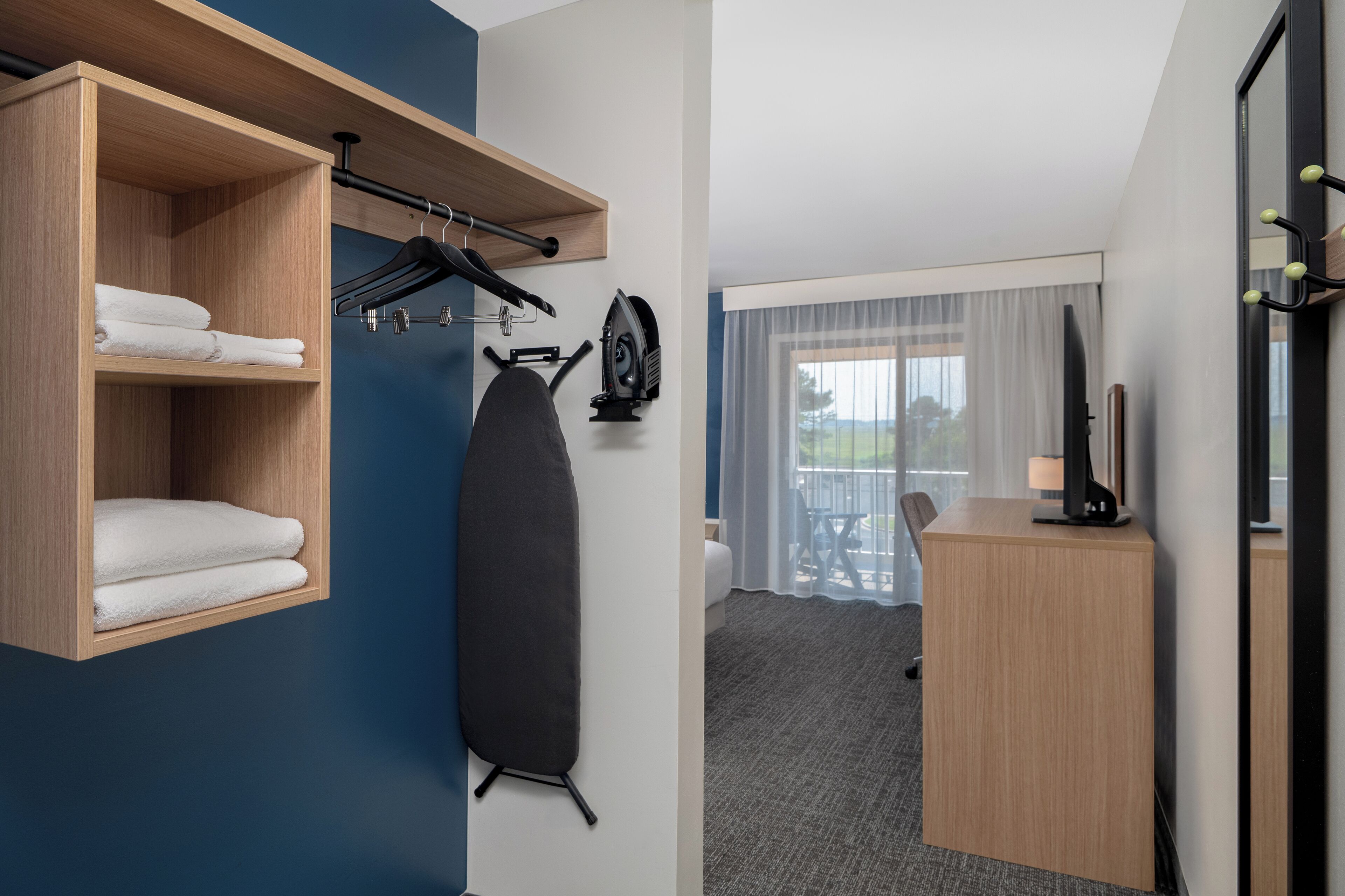 A hotel room with a wooden shelf holding towels, a black ironing board, and a desk with a lamp.