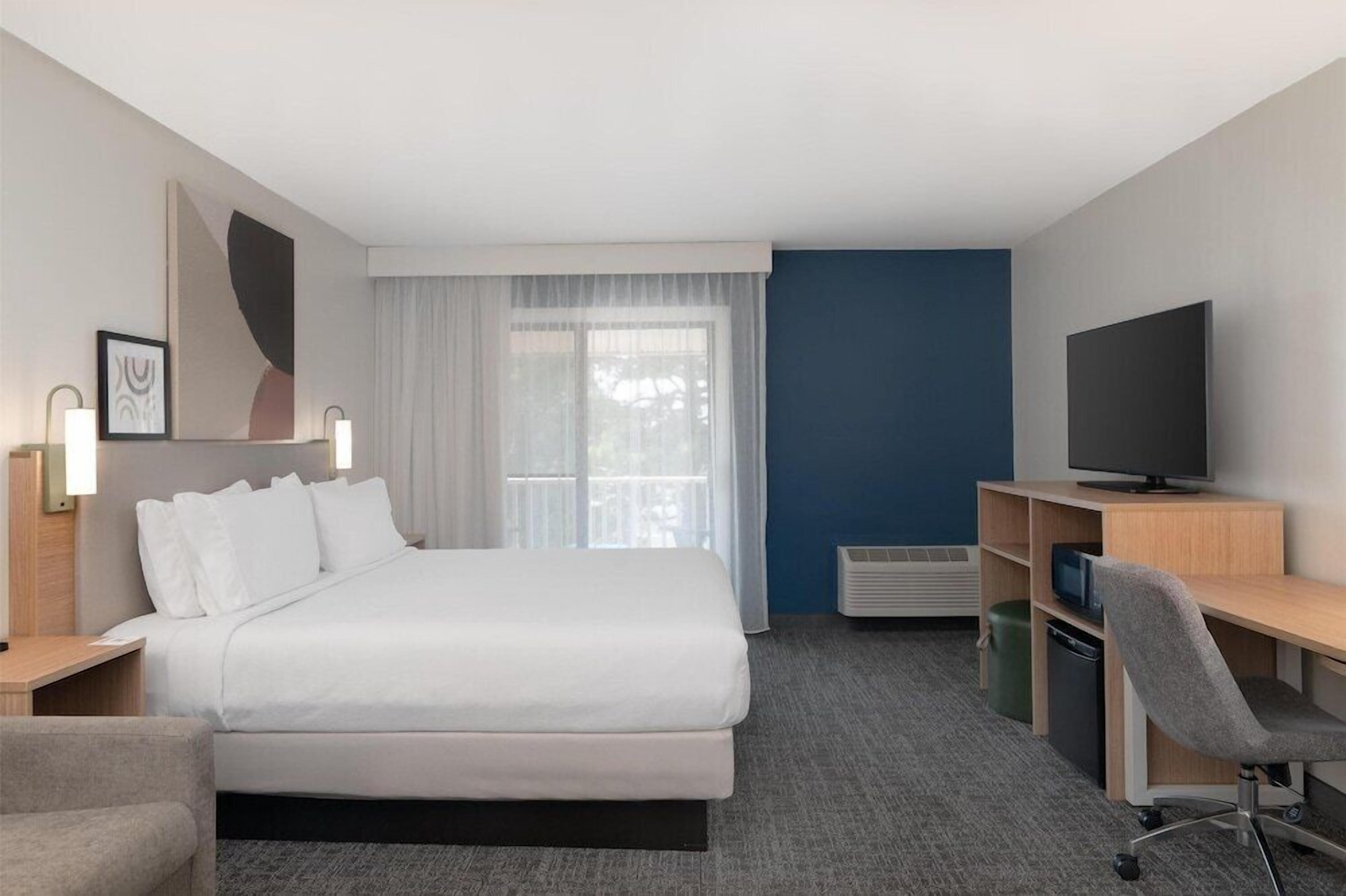 A hotel room with a bed, a desk with a chair, a television, and a window with curtains.