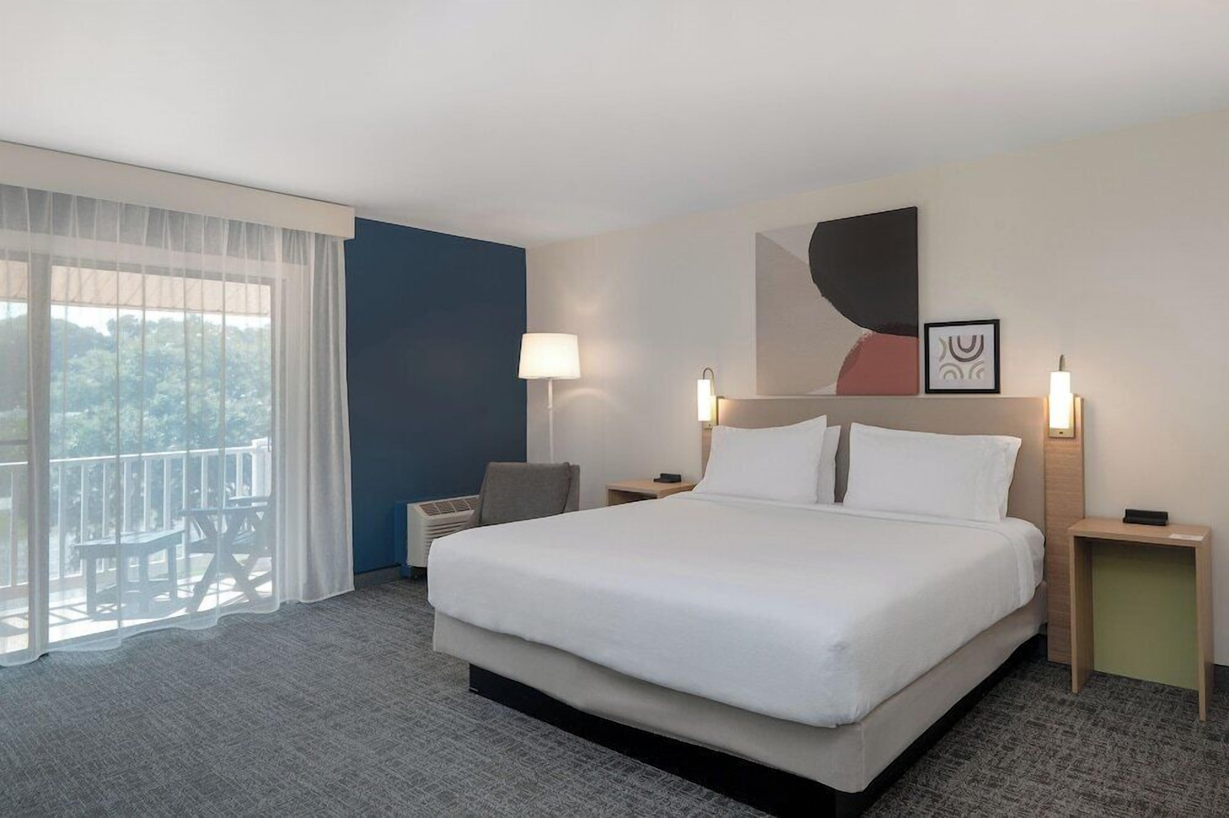 A hotel room with a large bed, two bedside tables, a chair, a balcony with a view, and a painting on the wall.