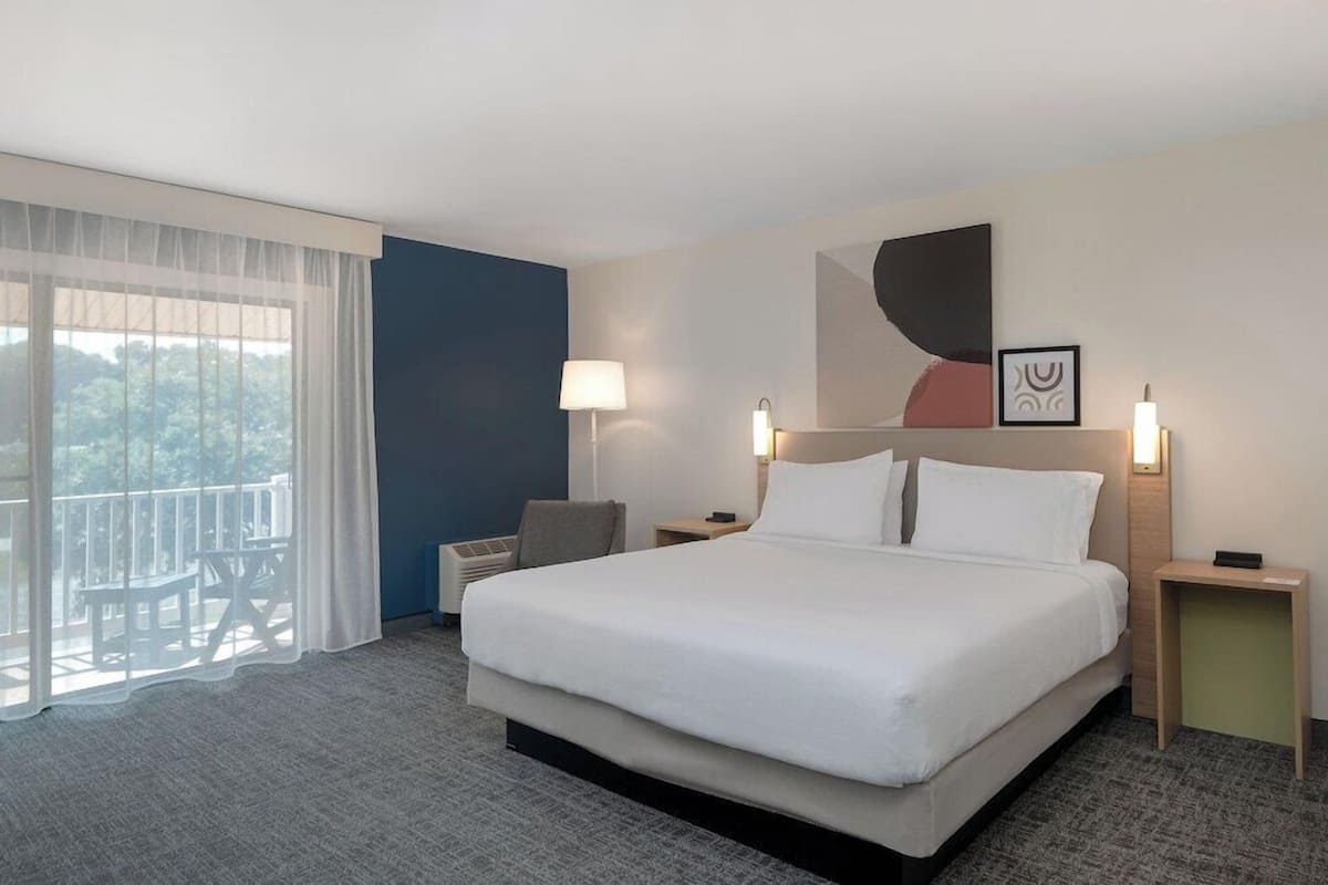 A hotel room with a large bed, two bedside tables, a chair, a balcony with a view, and a painting on the wall.