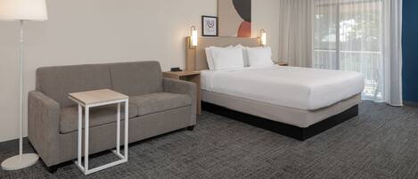 Habitación de hotel con cama, sofá, una mesita, un buró, lámpara y un cuadro en la pared.