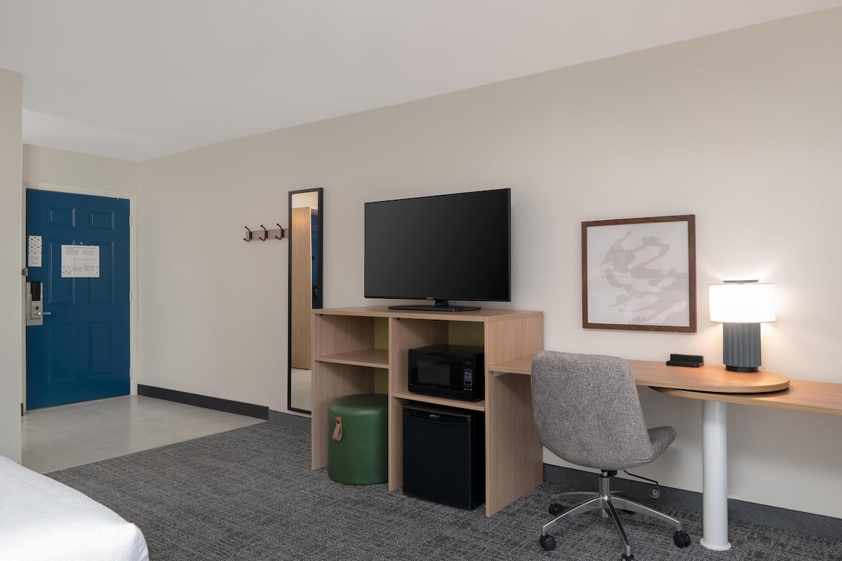 A hotel room with a bed, a desk with a TV, a microwave, a small fridge, a green trash bin, a grey office chair, a lamp, and a blue door.