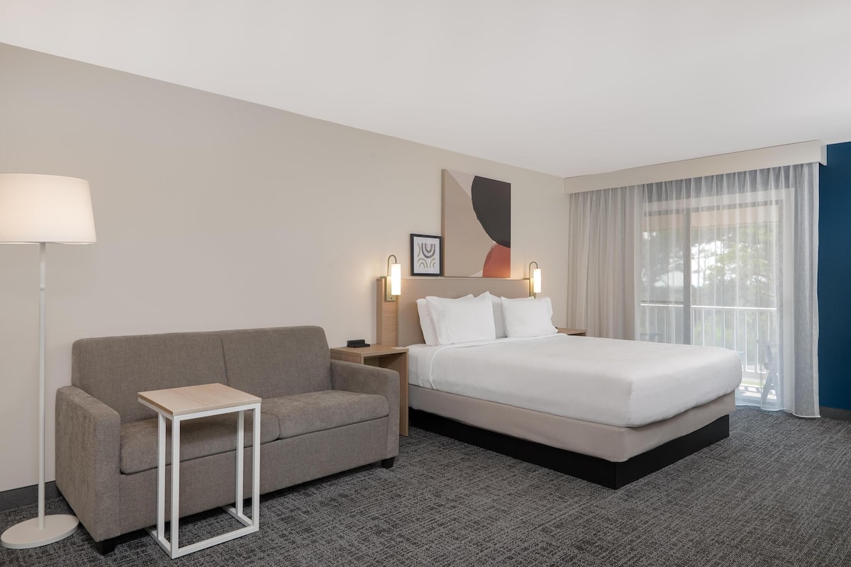 A hotel room with a bed, a sofa, a small table, a nightstand, a lamp, and a window with curtains.