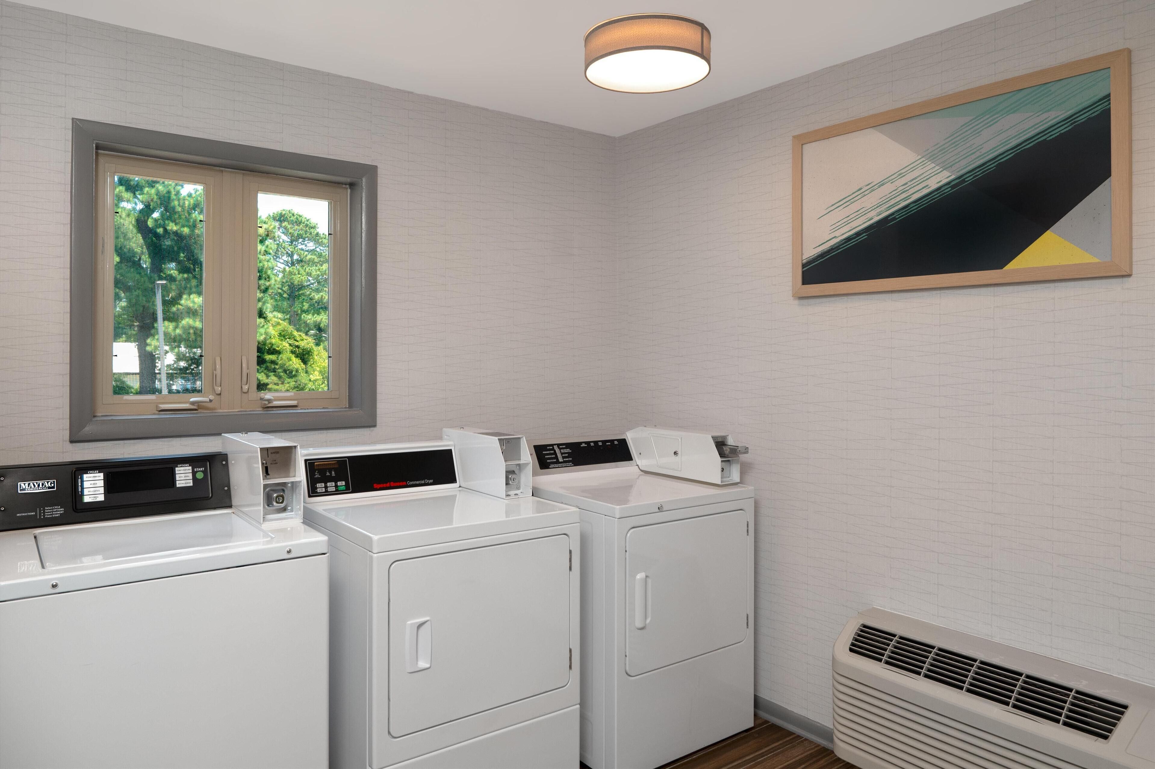 Une buanderie équipée de deux machines à laver, d’un sèche-linge, d’une bouche d’aération et d’une fenêtre donnant sur les arbres.
