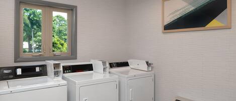 A laundry room with two washing machines, a dryer, a vent, and a window with a view of trees.