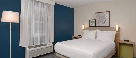 A hotel room with a bed, a standing lamp, a window with curtains, a wall-mounted artwork, and a bedside table with a lamp.