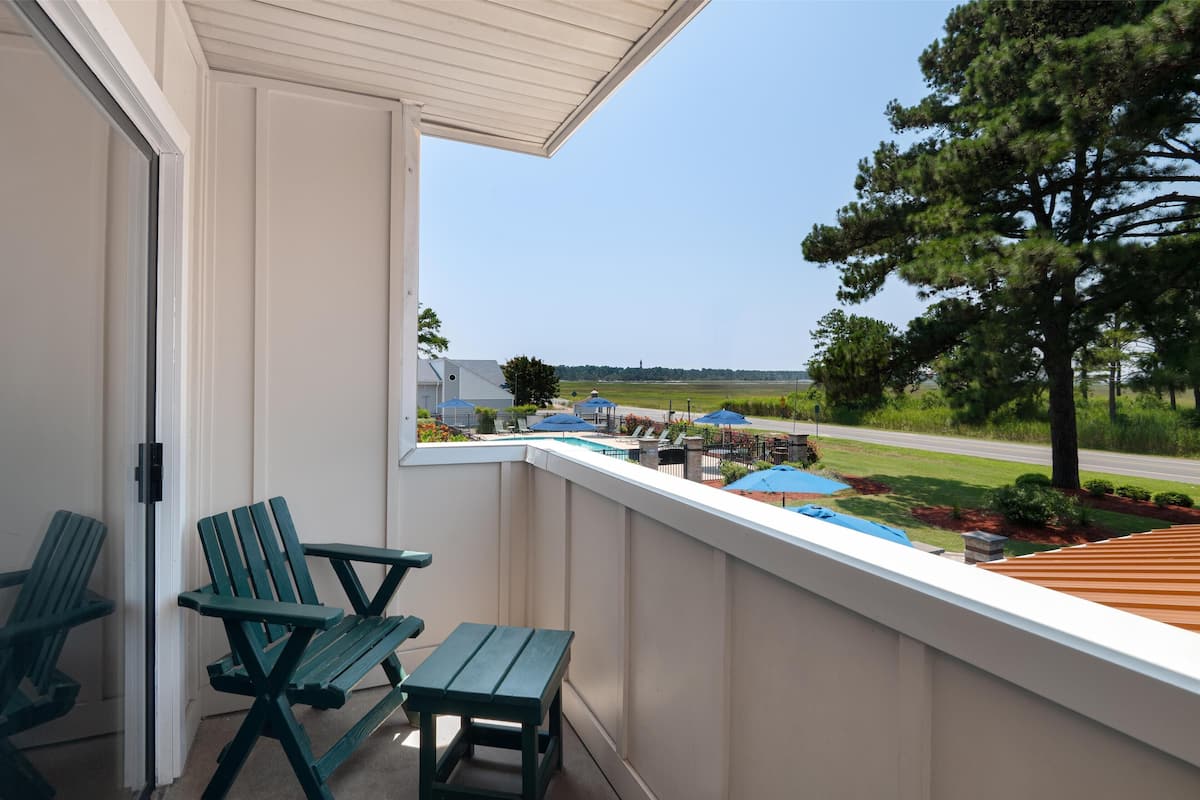 A balcony with two green Adirondack chairs and a small table, overlooking a landscaped area with umbrellas and a road.