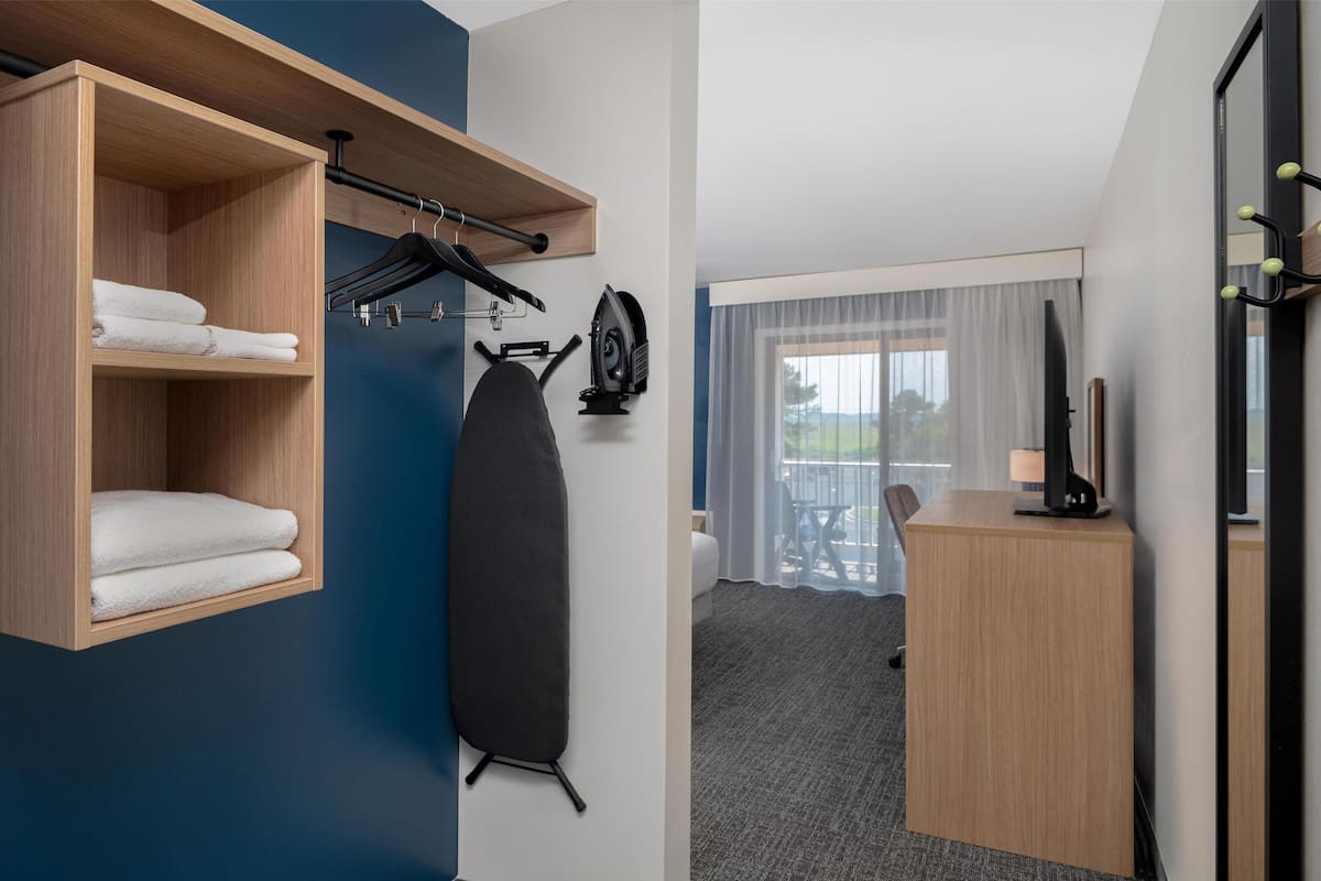 A hotel room with a wooden shelf holding towels, a black ironing board, and a desk with a lamp.