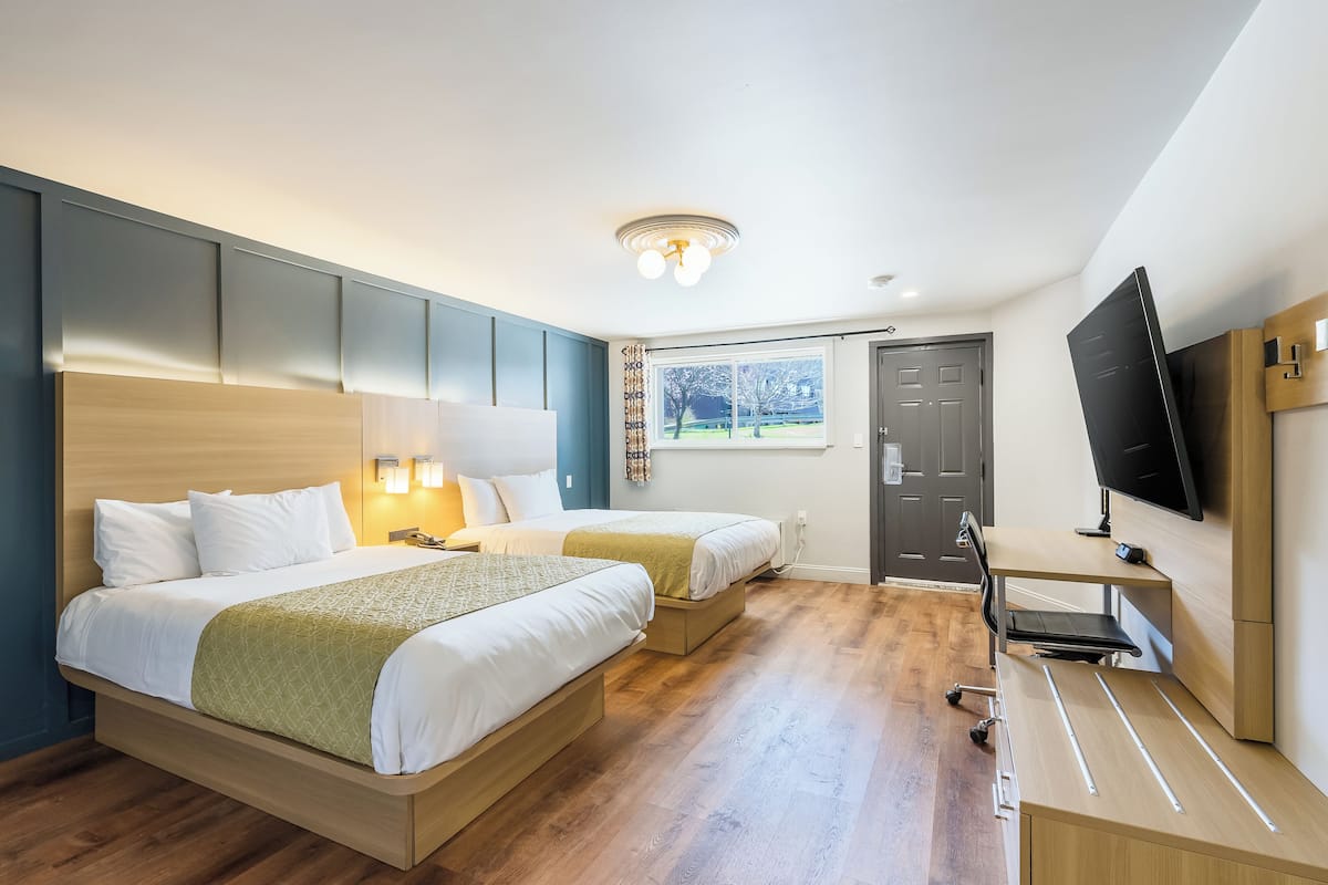A hotel room with two beds, a desk, a TV, and a window with a view of trees.