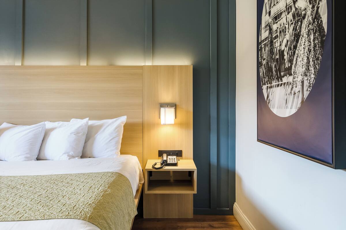 A neatly made bed with white linens and a beige throw blanket, a bedside table with a lamp, and a framed cityscape picture on the wall.
