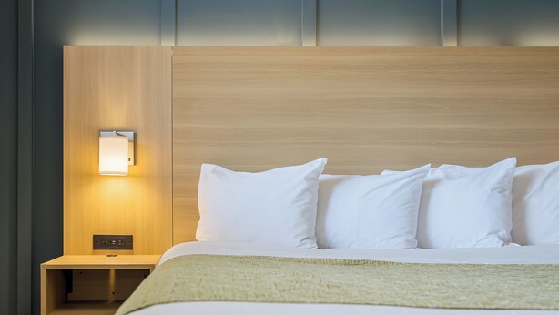 A hotel room with a wooden headboard, a bedside table with a lamp, and a bed with white bedding and pillows. - Ardsley Acres Hotel Westchester (Ardsley)
