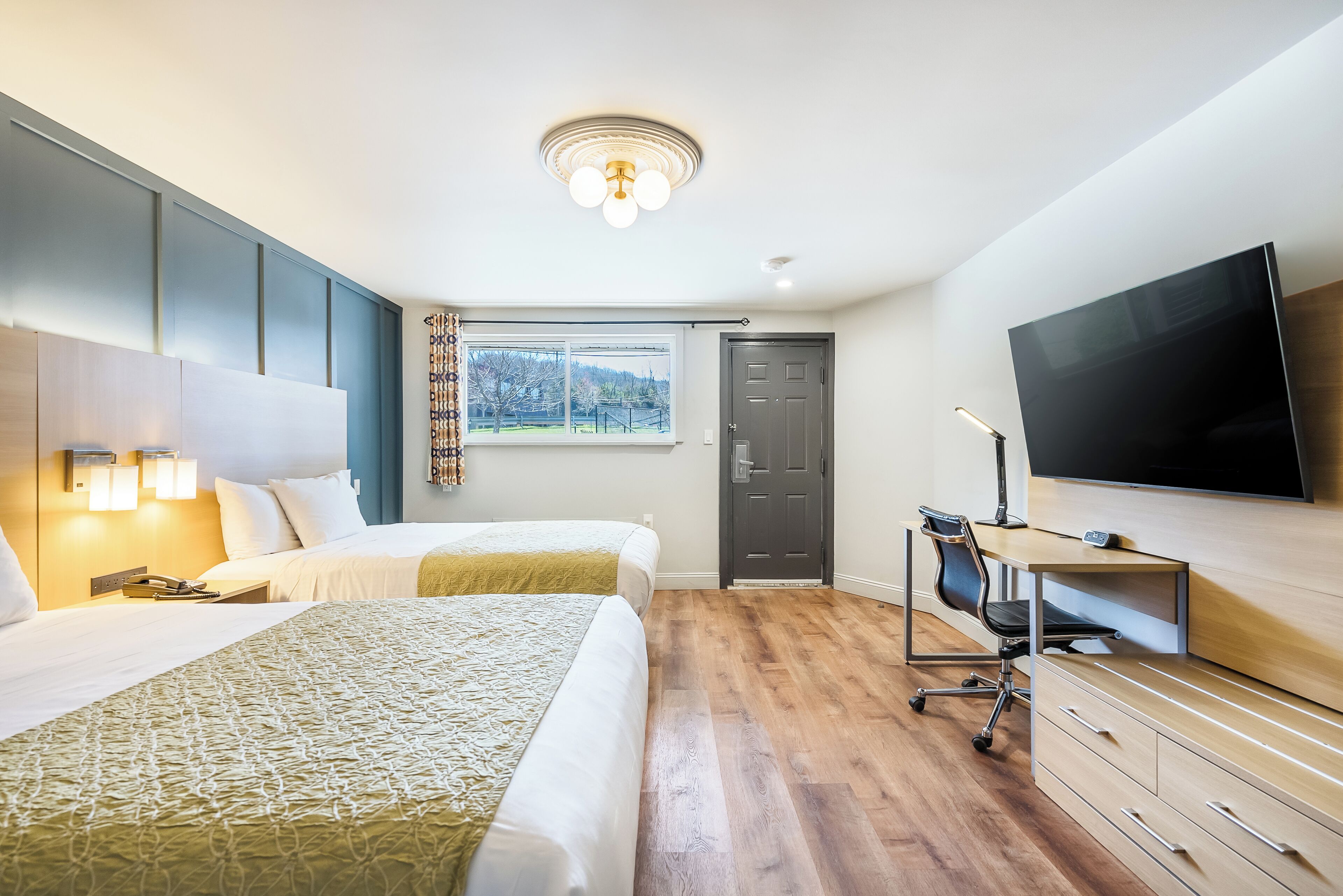 A hotel room with two beds, a desk with a chair, a flat-screen TV, and a window with a view of trees.
