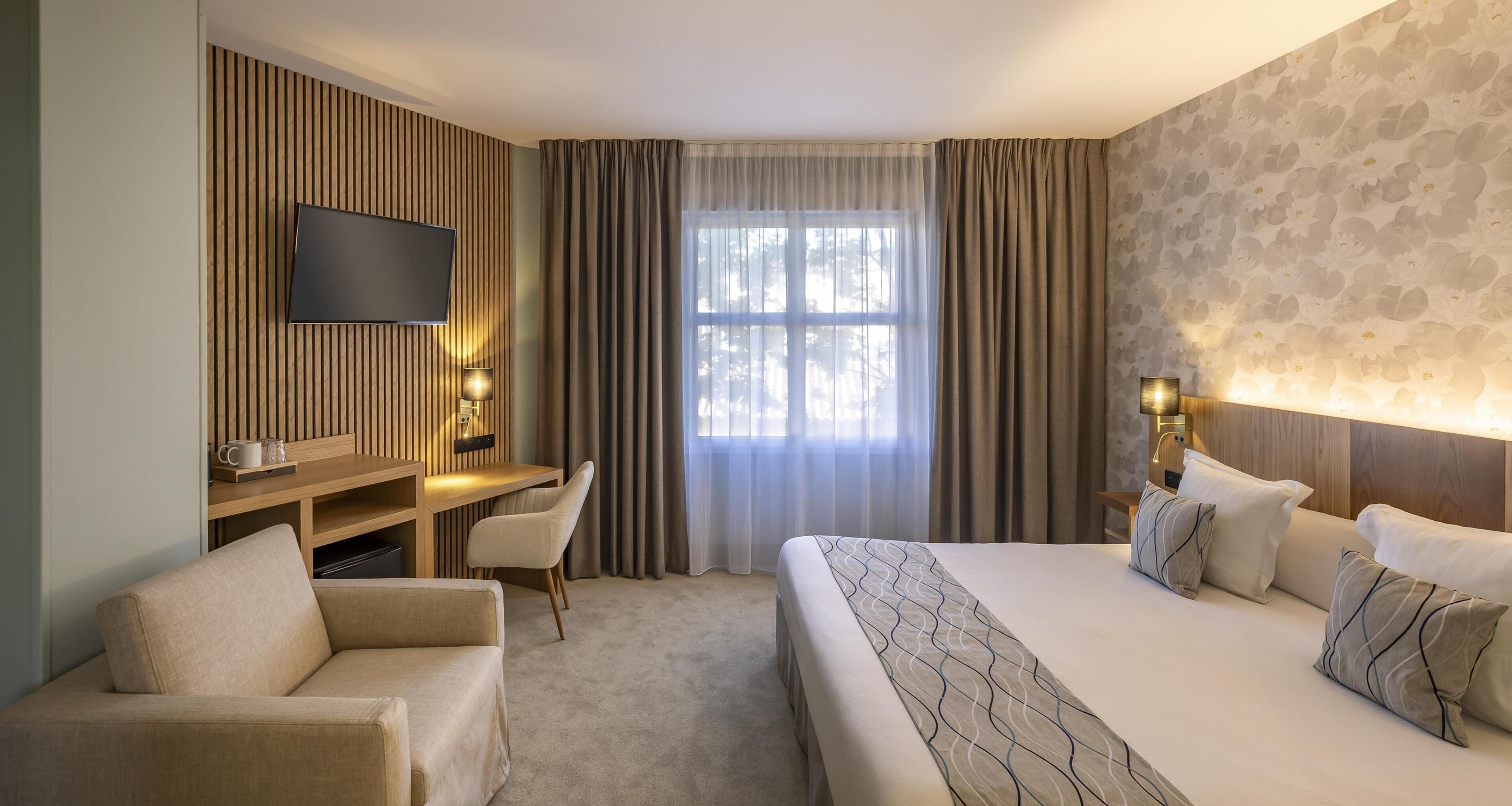 A hotel room with a bed, a desk, a chair, a TV, and a window with curtains.
