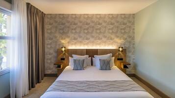 A hotel room with a large bed, two bedside tables with lamps, a window with curtains, and a patterned wallpaper.