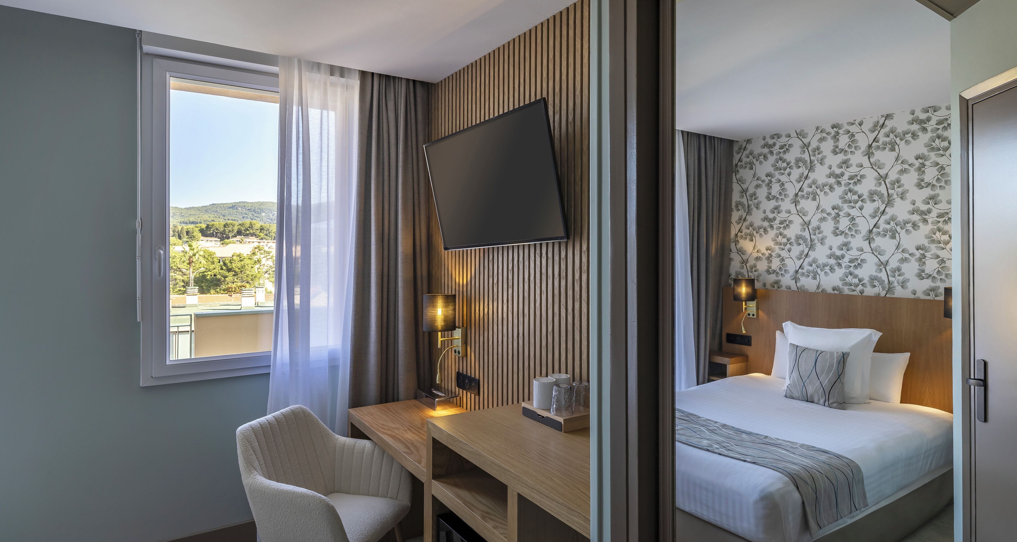 A hotel room with a bed, a desk with a chair, a TV mounted on the wall, and a window with a view of greenery.