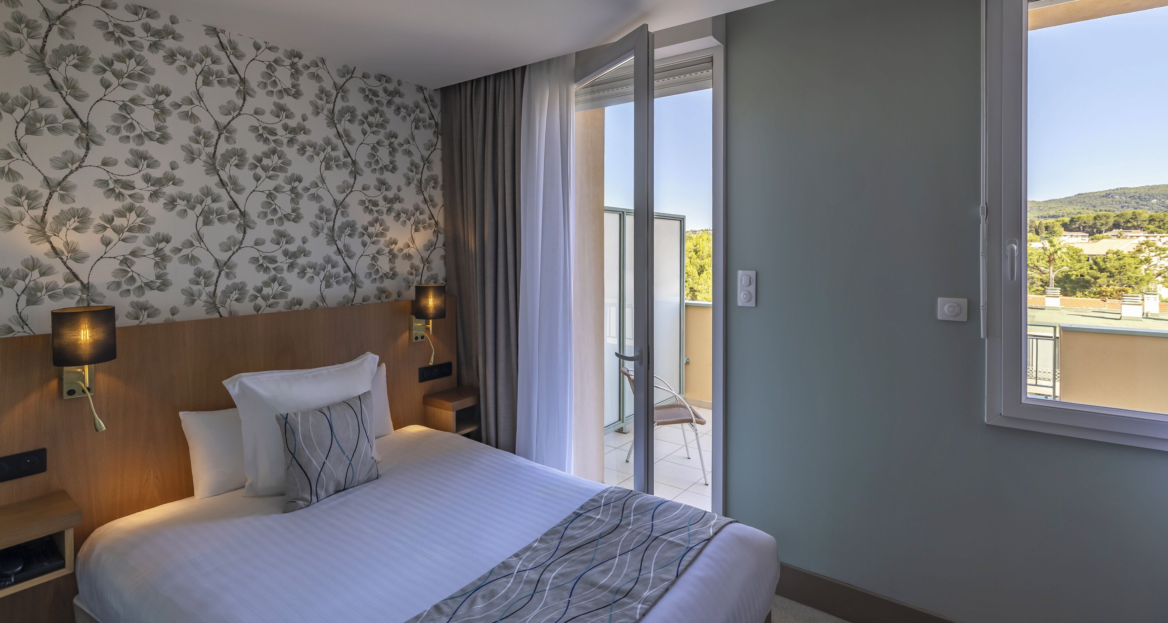 A hotel room with a bed, two wall-mounted lamps, a view of the outdoors through a sliding glass door, and a balcony with a chair.
