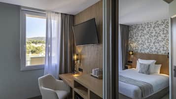 Quarto de hotel com cama, escrivaninha, cadeira, televisão e janela com vista para o exterior.