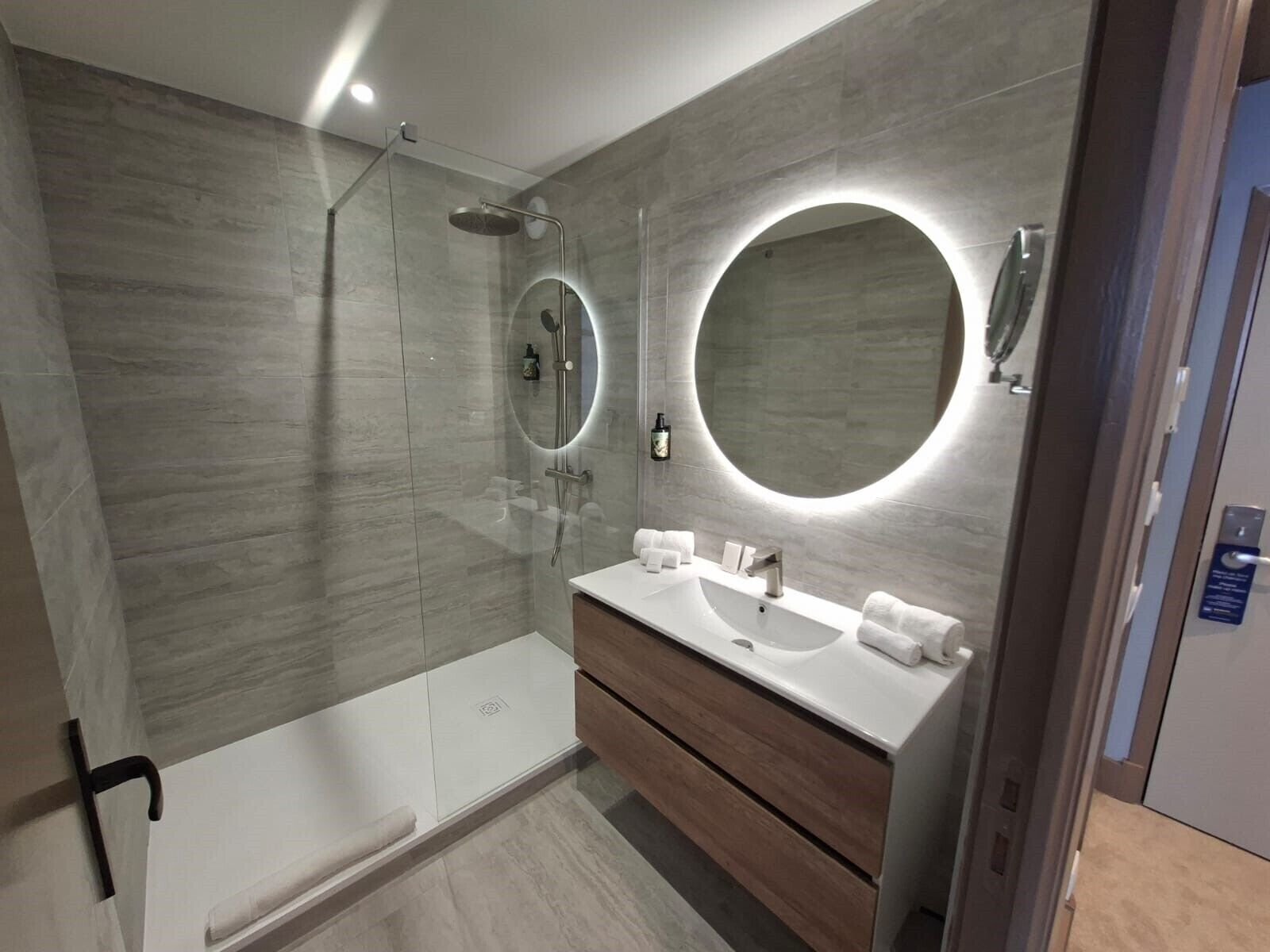 A modern bathroom with a glass-enclosed shower, a round mirror, and a vanity with a sink and towel rack.