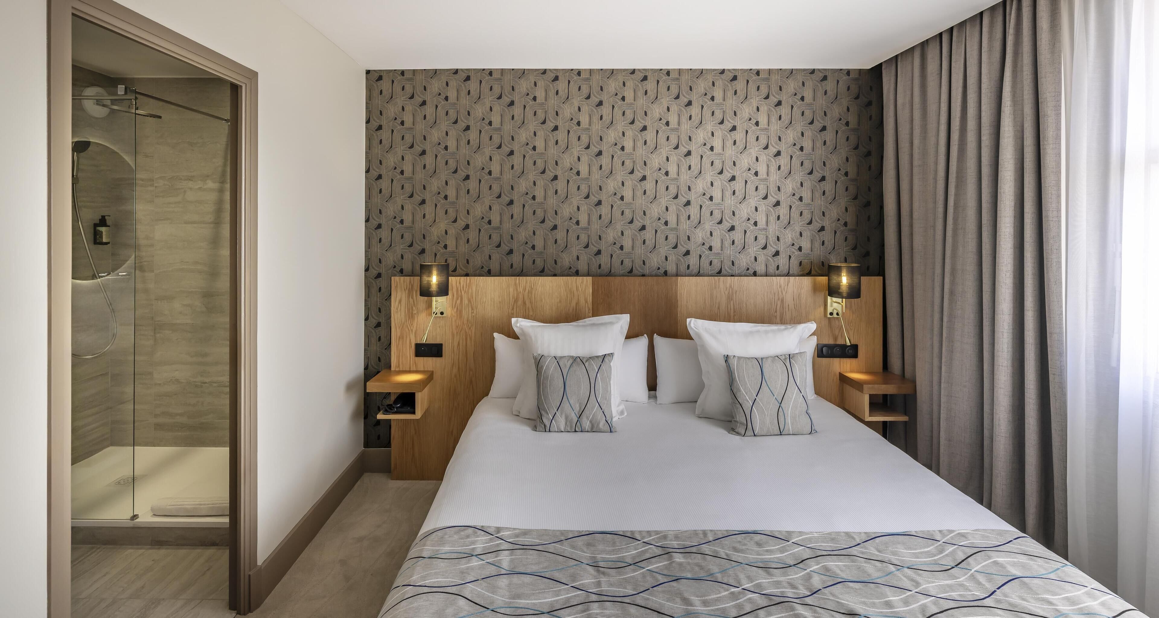 A hotel room with a large bed, a headboard with a geometric pattern, bedside lamps, and a shower area.