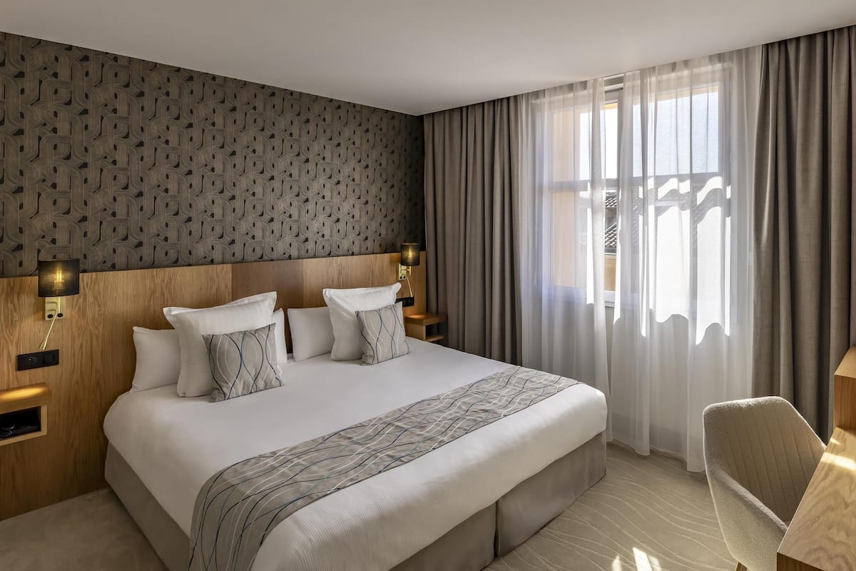 Habitación de hotel moderna con una cama grande, cabecera de madera, escritorio con lámpara y vistas al exterior a través de cortinas traslúcidas.