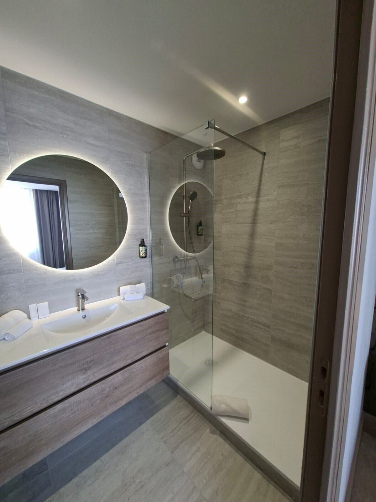 A modern bathroom with a glass-enclosed shower, a large mirror with a circular light, and a vanity with two sinks.