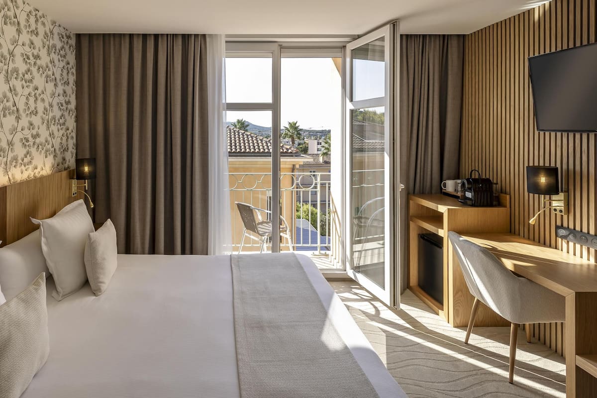 Habitación de hotel con una cama grande, un escritorio con silla, televisor y balcón con vistas a la ciudad.