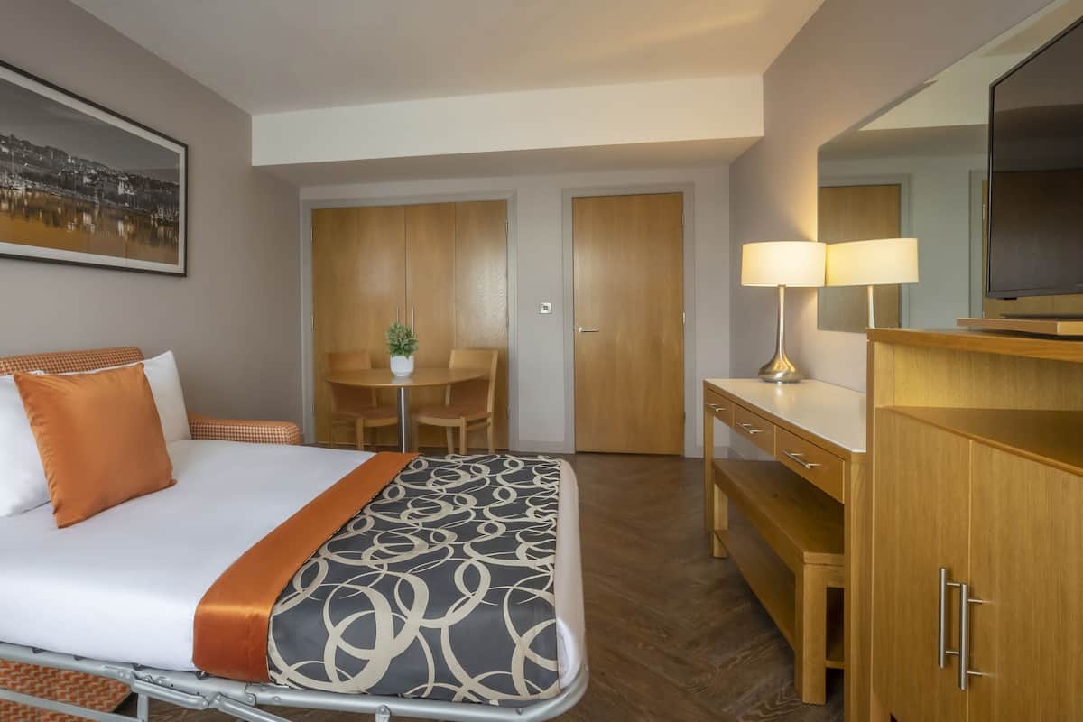 A hotel room with a bed, a desk, a chair, a lamp, and a television.