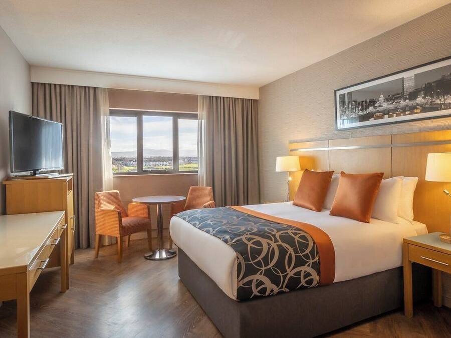 Une chambre d’hôtel comprenant un lit, deux tables de chevet, un bureau, une chaise, un téléviseur et une fenêtre avec des rideaux.