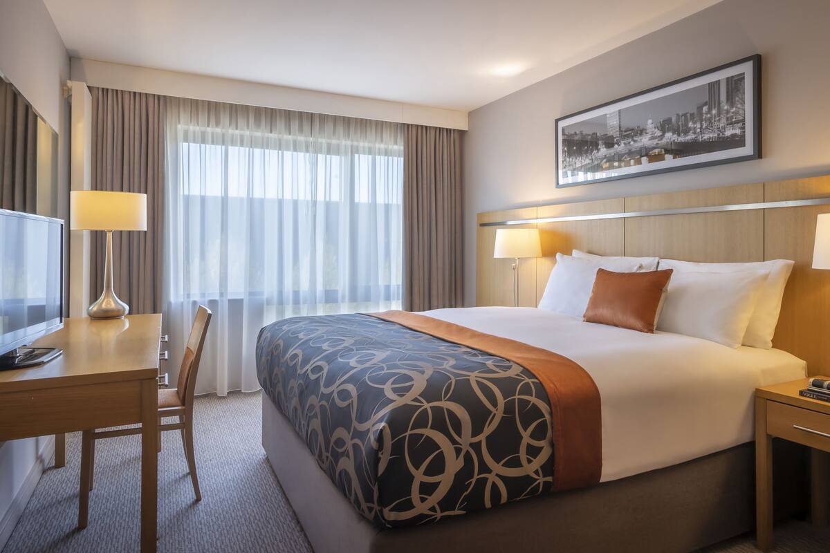 A hotel room with a large bed, two bedside lamps, a desk with a TV, and a chair.