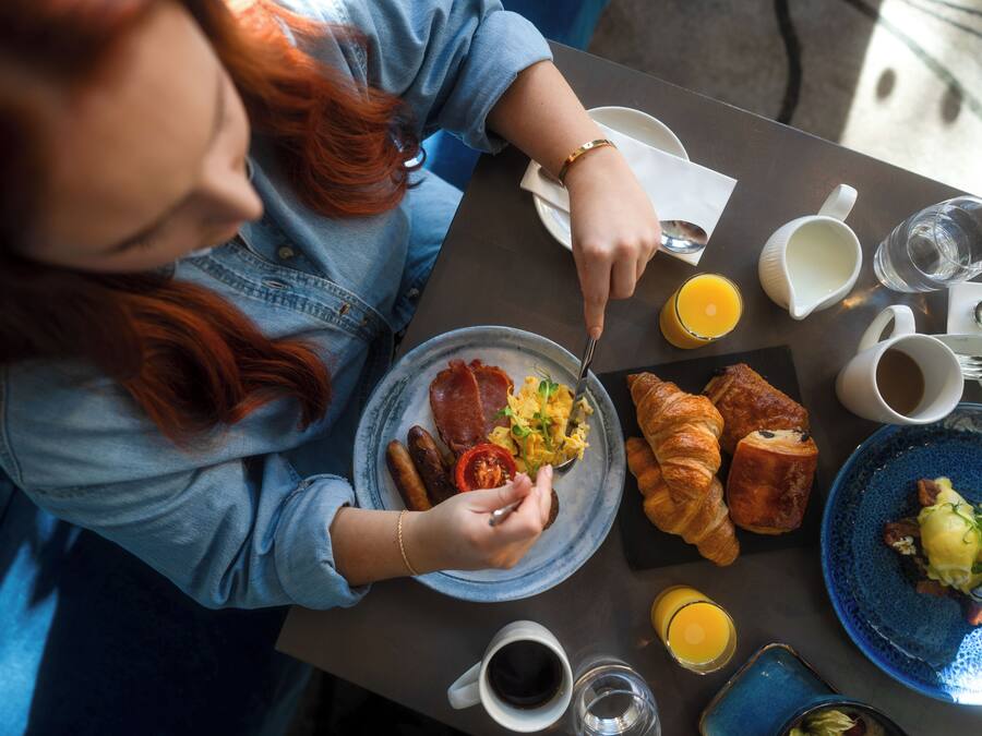 A person dining at a table with breakfast items including croissants, eggs, and a plate of sausages.