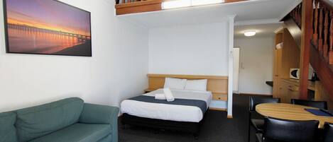 Habitación de hotel con cama, sofá verde, mesa de comedor con sillas y un cuadro con atardecer sobre un puente.