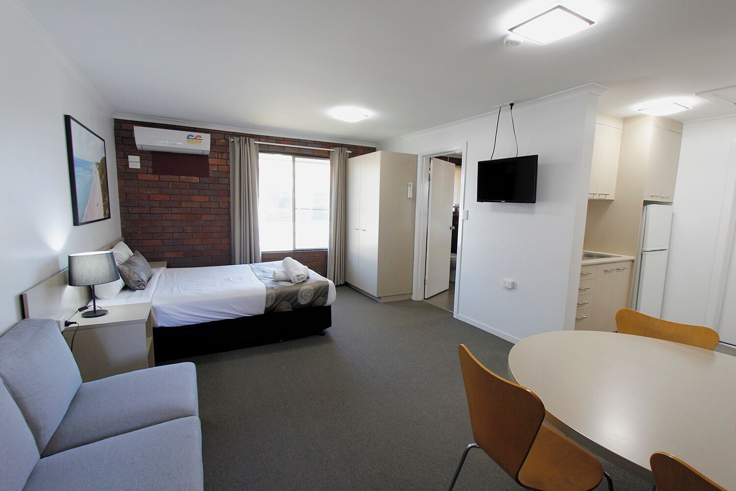 A compact hotel room with a bed, a desk, a chair, a TV, and a kitchenette.