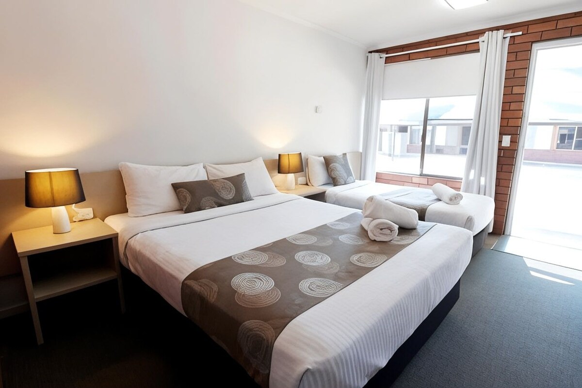 A hotel room with two beds, a nightstand with a lamp, and a view of the outside through a window.