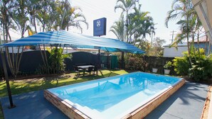 Une piscine rectangulaire bordée de briques, entourée d’une terrasse dotée d’un parasol bleu et blanc à rayures qui offre de l’ombre.