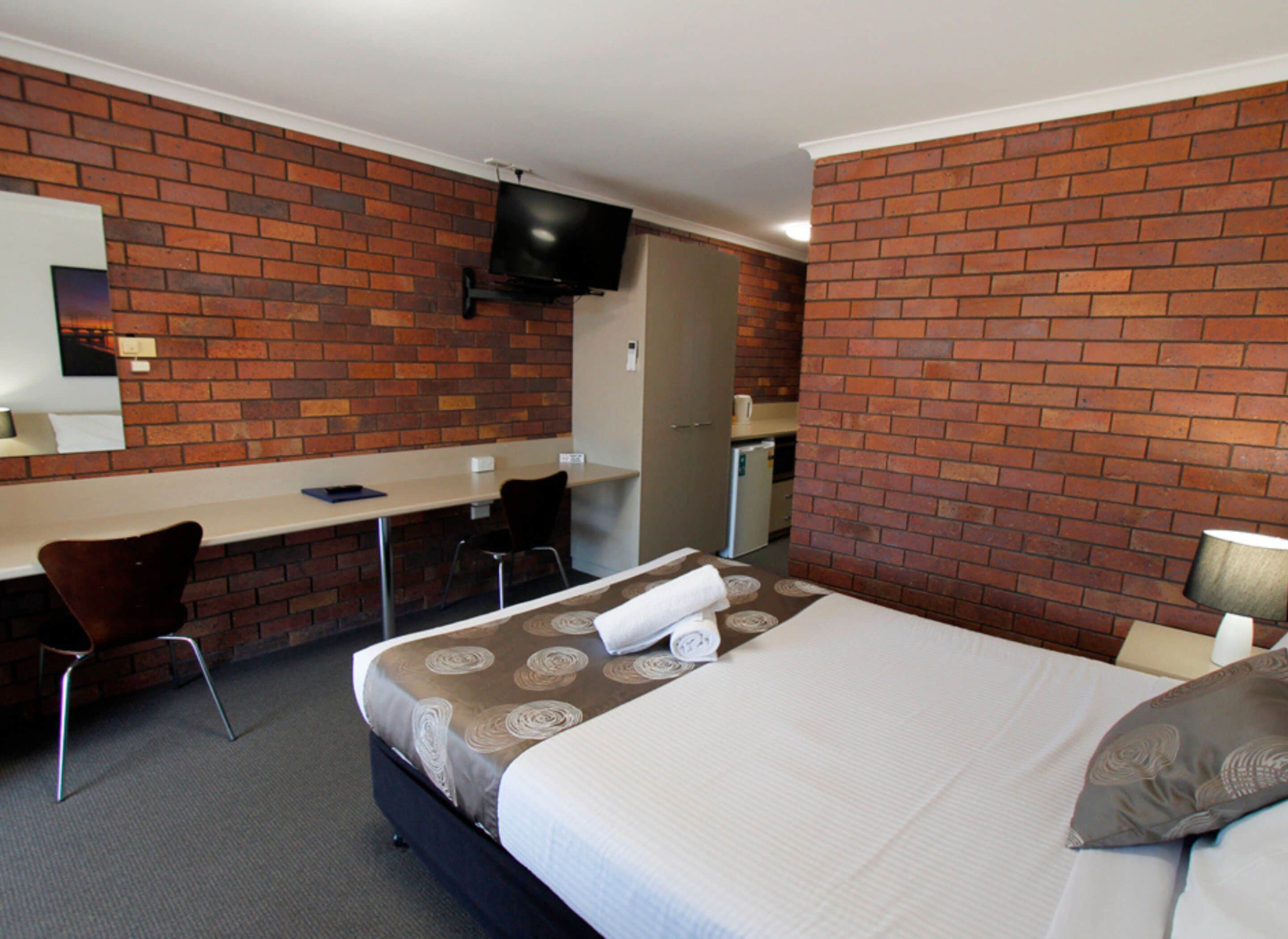 Une chambre d’hôtel avec un mur en briques, un lit recouvert d’une couvre-lit à motifs, un bureau avec une chaise, un téléviseur fixé au mur et une petite kitchenette.