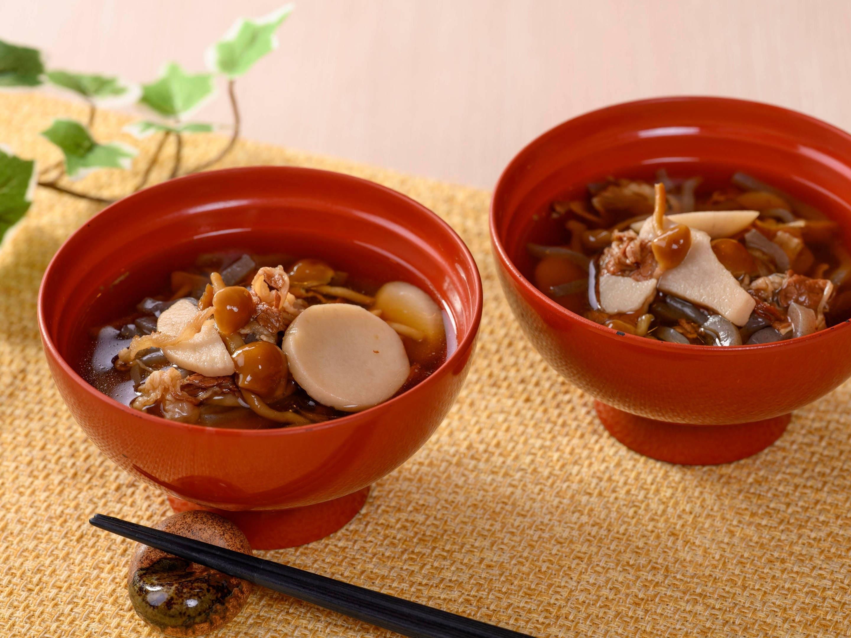 Two red bowls of soup with chopsticks and a woven mat.