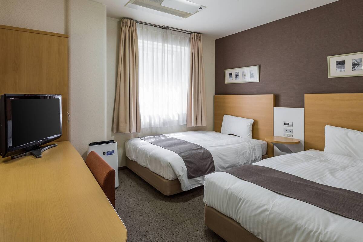 A hotel room with two beds, a desk, a television, and a window with curtains.