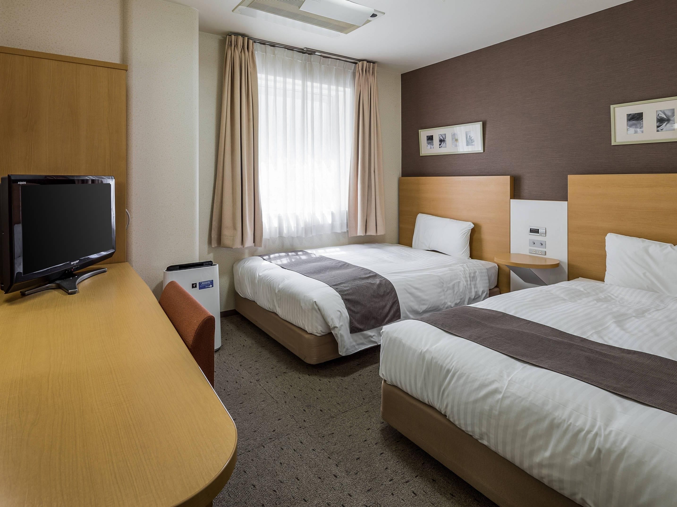 A hotel room with two beds, a desk, a television, and a window with curtains.