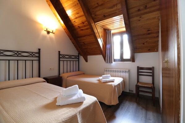 A room with two beds, wooden ceiling beams, a radiator, and a window with curtains.