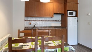 Cocina pequeña con armarios de madera, microondas y refrigerador. Hay una mesa de comedor con sillas y un mantel estampado.