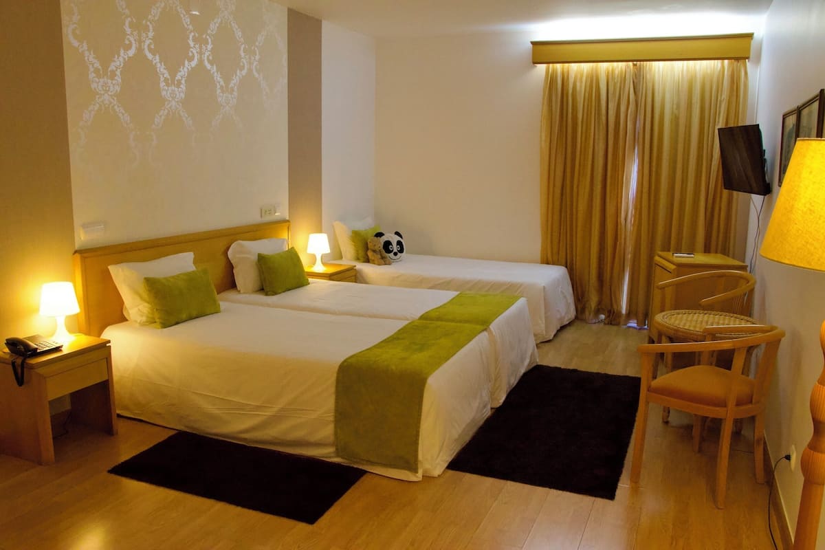Ein Hotelzimmer mit zwei Betten, einem Schreibtisch mit Lampe, einem Sessel, einer Panda-Kuscheltierfigur und einem an der Wand montierten Fernseher.